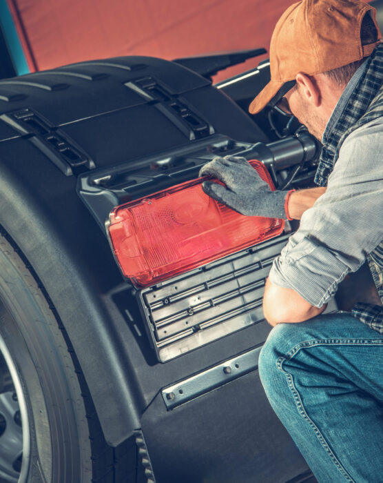 A truck mechanic works diligently on a truck's rear light in a well-lit workshop. The environment is busy, with tools and materials organized around him as he focuses on ensuring the safety of the vehicle.